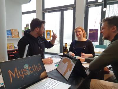 Image of a demo session of Mystika. A Narrator and two players are sitting at a table dressed with black table cloth. They all have character sheets and twelve sided dice laying in front of them. The narrator is explaining a situation, pointing a finger to support his narration. Two players listen to his explanation, reacting amused. In the foreground, a Mystika logo is displayed on a screen. Another screen displays a rough map of Amber City. The wall in the background consists of tiled windows showing heavy fog outside.